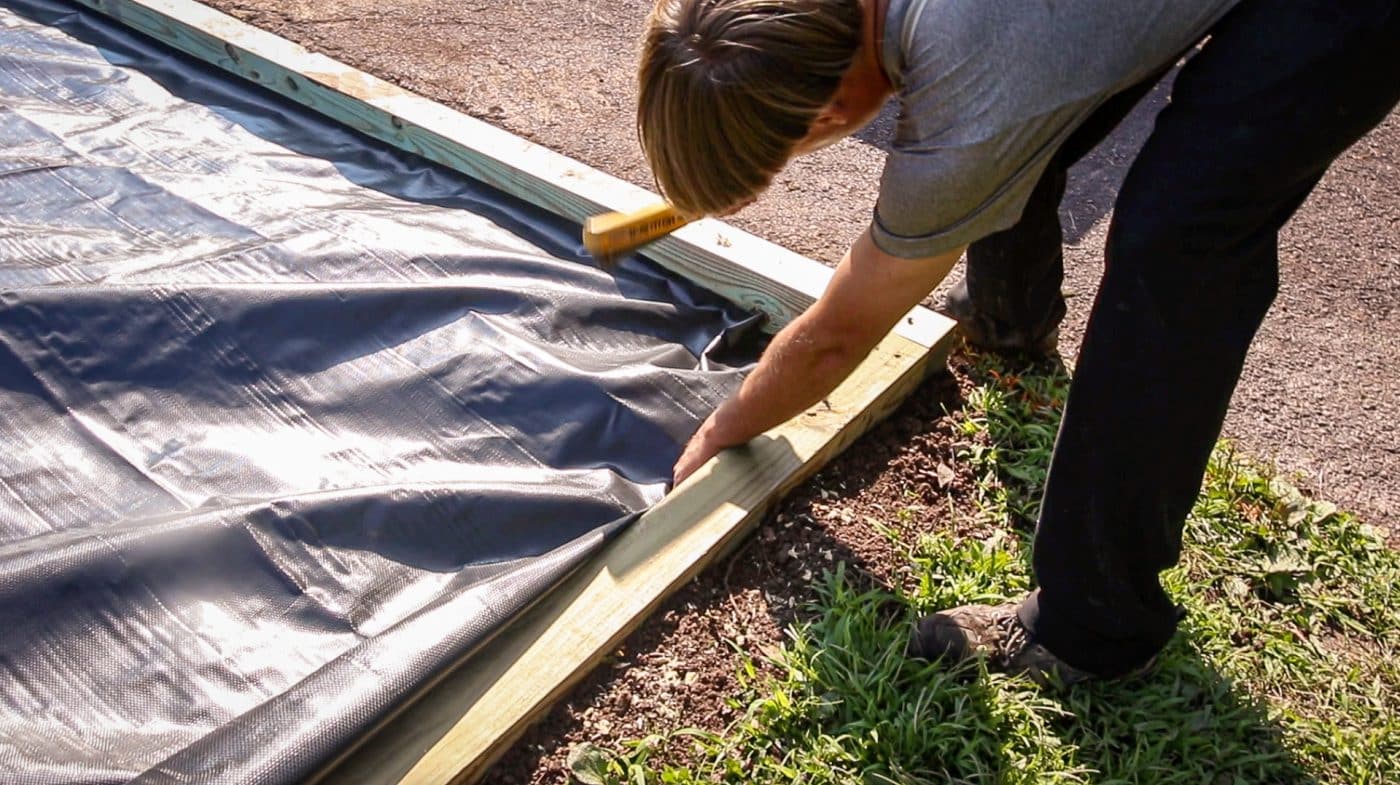 worker stapling the weed fabric to the perimeter as a step for how to build a shed base on uneven ground